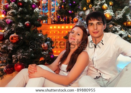 Young man and woman wearing white shirts sit near green trees in snow with Christmas balls