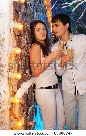 Young man and woman wearing white shirts with glasses of champagne stand near stack of wood