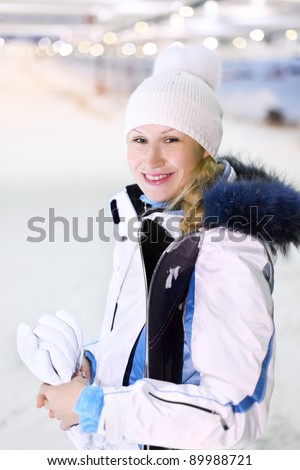 Young happy woman dressed in white sports clothes stands with skis and mittens in hands in indoor ski