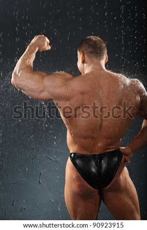 Undressed bodybuilder stands in rain back to camera and shows muscles of hand