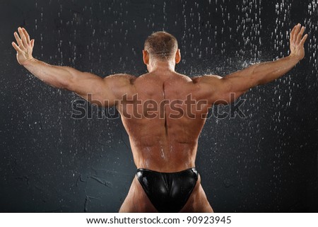 Undressed bodybuilder stands in rain back to camera and his arms in side