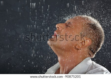 Tanned man wearing white wet shirt stands in rain and drops fall on his head.