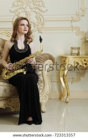 Pretty woman with sax sits on armchair in baroque studio with molding on walls