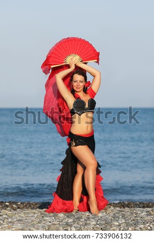 Dancer woman in black and red suit with fan dancing on seashore, fan overhead