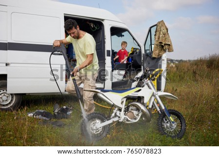Man washes motorbike after training outdoor near minibus, little son looks at him from driver cabin.