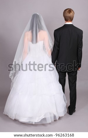 Groom and beautiful bride stand their backs to camera in studio on gray background