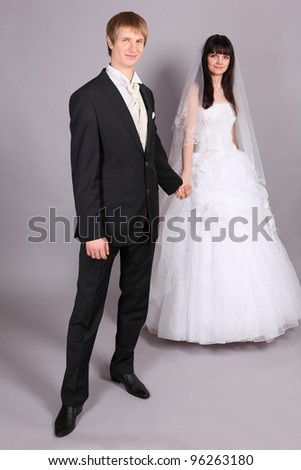 Beautiful bride and groom hold hands and stand in studio on gray background; bride stands behind groom; focus on man