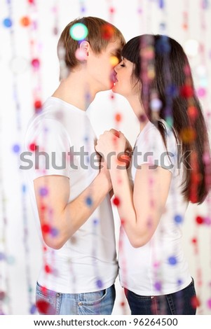 Man and woman dressed in white shirts hold hands and kiss behind transparent curtain of beads; focus on pair
