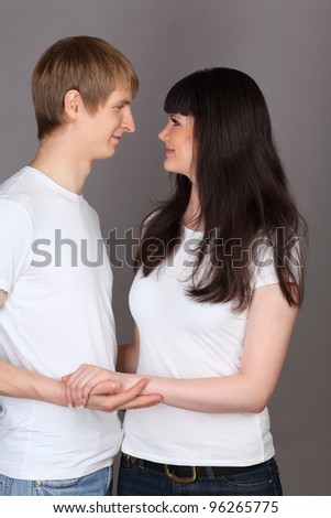 Happy man and woman dressed in white shirts holding hands and looking at each other on gray background