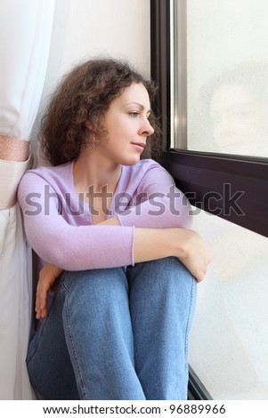 Young beautiful woman sits on windowsill, looks out window and dreams