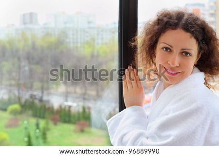 Smiling woman dressed in white bathrobe stands near window and looks into camera