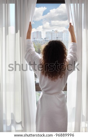 Woman dressed in white bathrobe opens curtains and looks up; woman stands back to camera