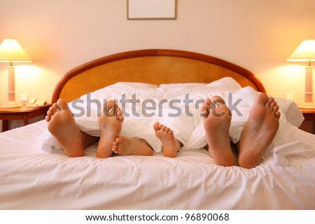 Mother, father and child rest on soft big bed with white sheets; focus on feet