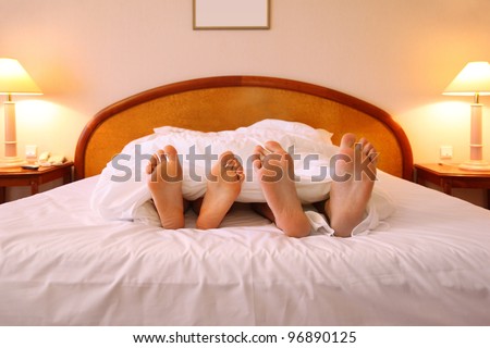 Woman and man relaxing on soft big bed with white sheets; focus on feet