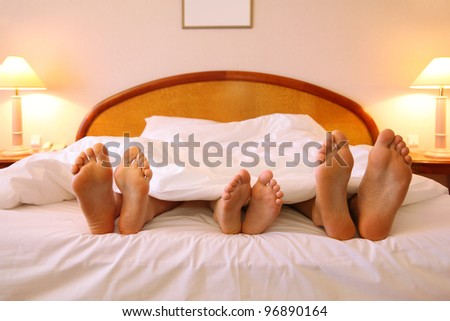 Mother, father and child lie on soft bed with white sheets; focus on feet