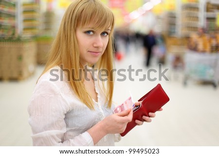 Blonde girl gets money from her red purse in store and looks at camera; shallow depth of field