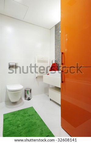 Modern, mostly white bathroom with colorful orange, red, and green accents. Modern toilet and sink basin with a fun bright green rug and bright orange cabinet doors.