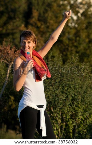 A young beautiful woman feeling victorious after a successful run in the park. She's wearing  black and white athletic clothing, has a towel around her neck and a water bottle in her right hand.