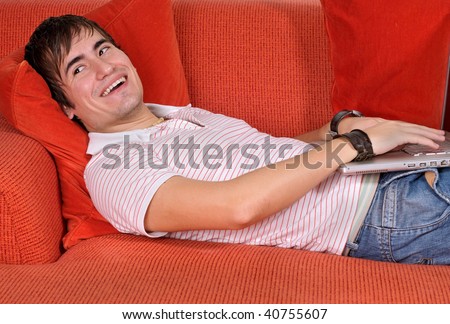 A young man relaxing with his laptop computer, while laying on the sofa, looking back and smiling.