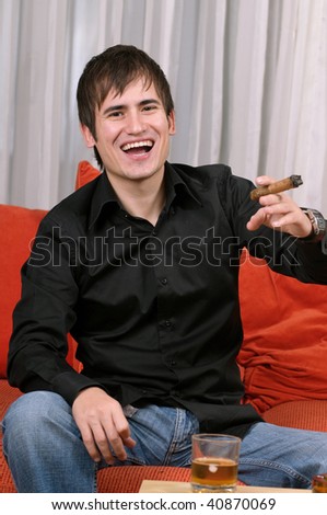 A man seated on a couch smiling with a cigar in one hand and a scotch in front of him.