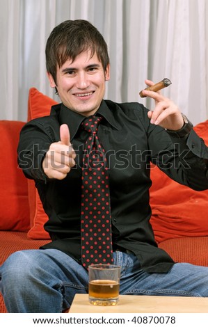 A young man seated on a bright orange sofa, wearing a black button down shirt smiling as he holds a cigar in his hand. There is a drink in front of him.