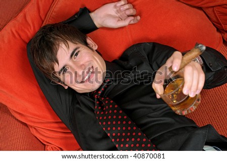 A young man laying on his orange couch with a drink and cigar in his hand. Photo taken from above.