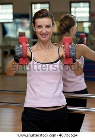 A young woman working out with red dumbells at a gym in front of a mirror.