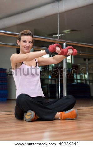 A focused pretty young woman exercising with dumbells, seated on a wooden gym floor in front of a glass wall.