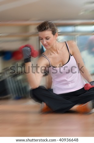 A focused pretty young woman exercising with dumbells, seated on a wooden gym floor in front of a glass wall.