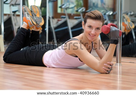 A beautiful young smiling woman laying on her stomach on a wooden gym floor in front of a mirror exercising with dumbell weights.
