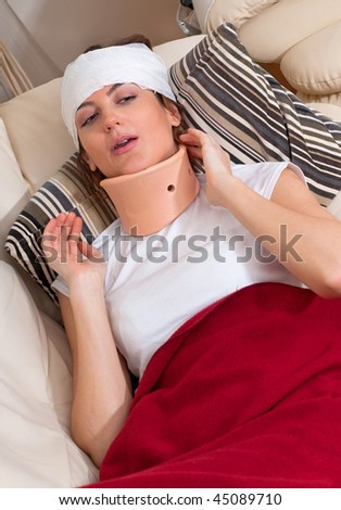A woman with a bandaged head and neck brace lays on the couch in pain.
