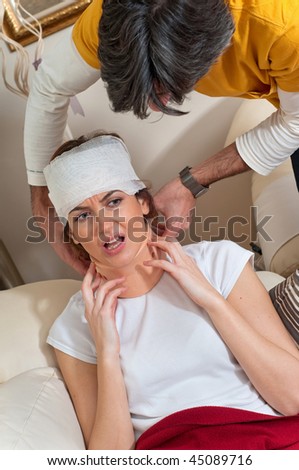 A man helps an injured woman wearing a neck brace and with bandages on her head as she tries to get comfortable. He is trying to adjust her neck brace to make her more comfortable.
