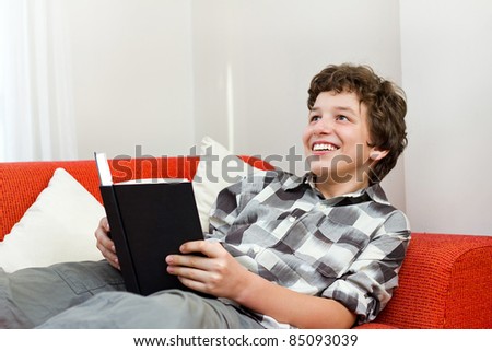 A preteen boy laying back on an orange couch with white pillows as he holds a black book in his hand and looks up with a big smile.