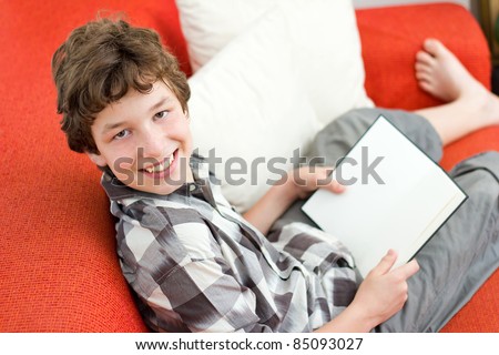 A preteen boy lounging on an orange couch with white pillows and reading a blank book looks up at the camera and smiles to show he enjoys reading.