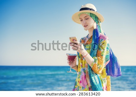 beautiful young woman is sms texting using an app on her smartphone, holding a martini cocktail on the beach