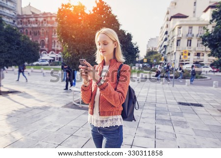 beautiful woman is sending a text message using an app on her smartphone while walking in the street on a sunset background