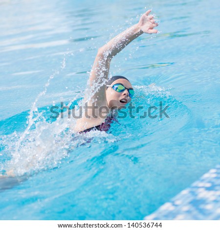 Young girl in goggles and cap swimming front crawl stroke style in the blue water pool