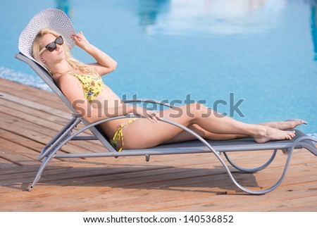 Young woman in bikini, sunglasses,  hat and swimsuit laying on chaise-longue and sunbathing by the pool in a summer vacation