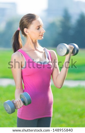 Portrait of cheerful woman in fitness wear exercising with dumbbell at green city park. Outdoors sport.