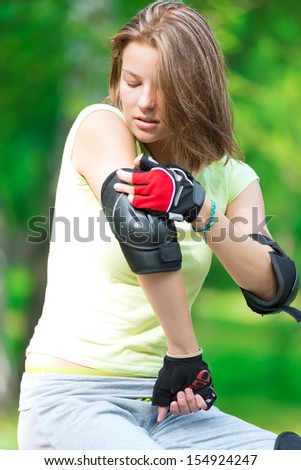 Woman skating in park. Girl going rollerblading sitting on bench  putting on elbow guard protection. Sporty caucasian woman in outdoor fitness activities.