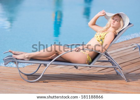 Young woman in bikini, hat and  swimsuit laying on chaise-longue and sunbathing by the pool in a summer vacation