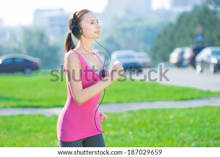 Jogging woman running in city park in sunshine on beautiful summer day and listening a music in headphones. Sport fitness model caucasian ethnicity training outdoor for marathon.