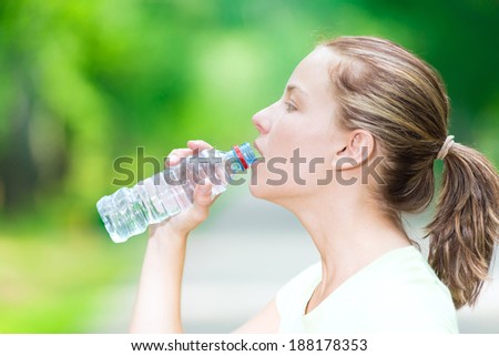 Young beautiful woman drinking cold mineral water from a bottle  after sport fitness exercise at green summer park