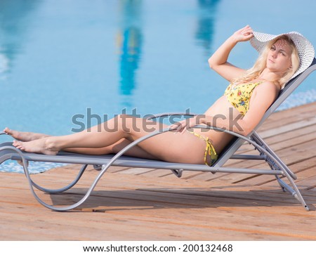 Young woman in bikini, hat and  swimsuit laying on chaise-longue and sunbathing by the pool in a summer vacation
