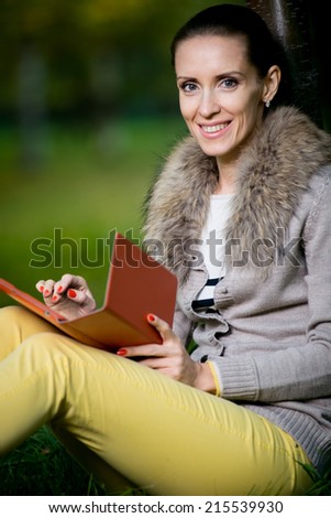 Fashion woman using a tablet computer outside in evening park. Student sitting on green grass over tree.