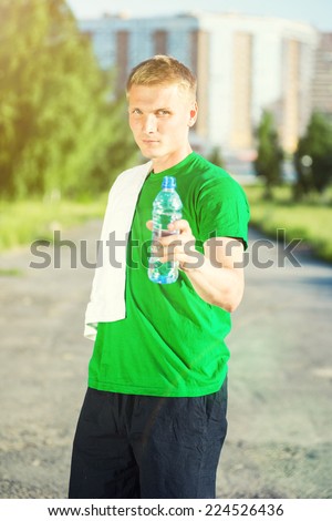 Tired man with white towel drinking water from a plastic bottle after fitness time and exercising in city street park at beautiful summer day. Sporty model caucasian ethnicity training outdoor.