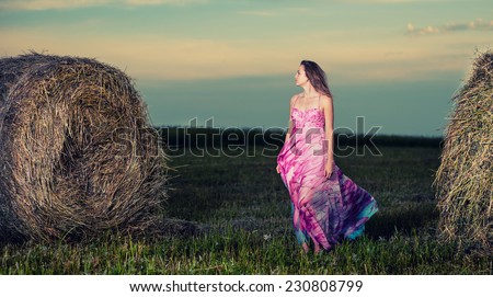 Young lady standing in evening field. Beautiful woman posing at the old rural farm location. Outdoor summer portrait of pretty fashion style woman in colored dress over haystack. Beautiful slim girl.