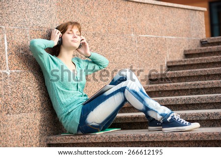 Young woman listening music in Headphones. Beautiful young female student.  Woman sitting on stairs in city park.
