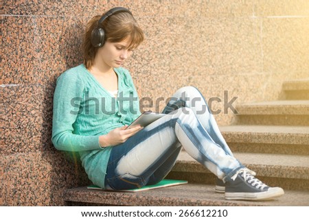 Preparing to exams outdoors. Headphones music. Beautiful young female student writing or reading something from note pad. Woman sitting on stairs in city park.