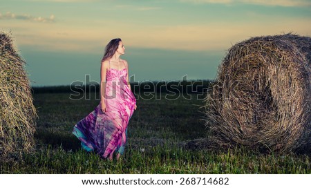 Young lady standing in evening field. Beautiful woman posing at the old rural farm location. Outdoor summer portrait of pretty fashion style woman in colored dress over haystack. Beautiful slim girl